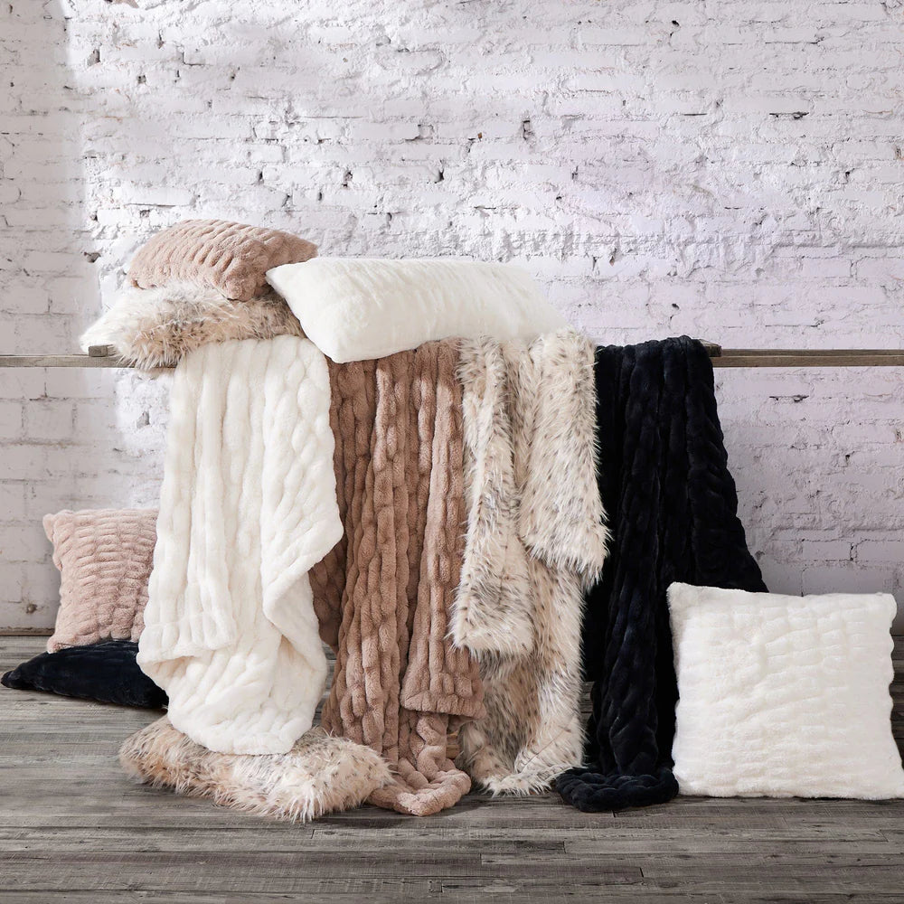 Collection of fluffy blankets and pillows on a wooden surface with a white brick wall background.