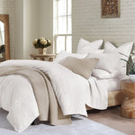 Neatly made bed with white and beige bedding in a bright bedroom.