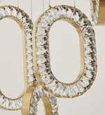 A gold-colored LED chandelier with a modern design, featuring circular and oval shapes with crystal accents, suspended by adjustable cords.
