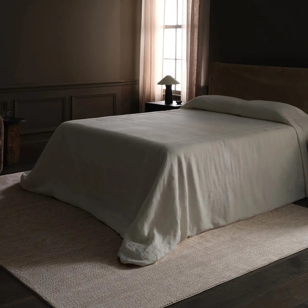 Bed with a white comforter in a dimly lit room with a lamp and curtains.