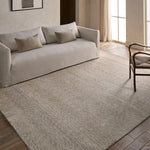Beige sofa with cushions on a textured rug in a room with large windows.