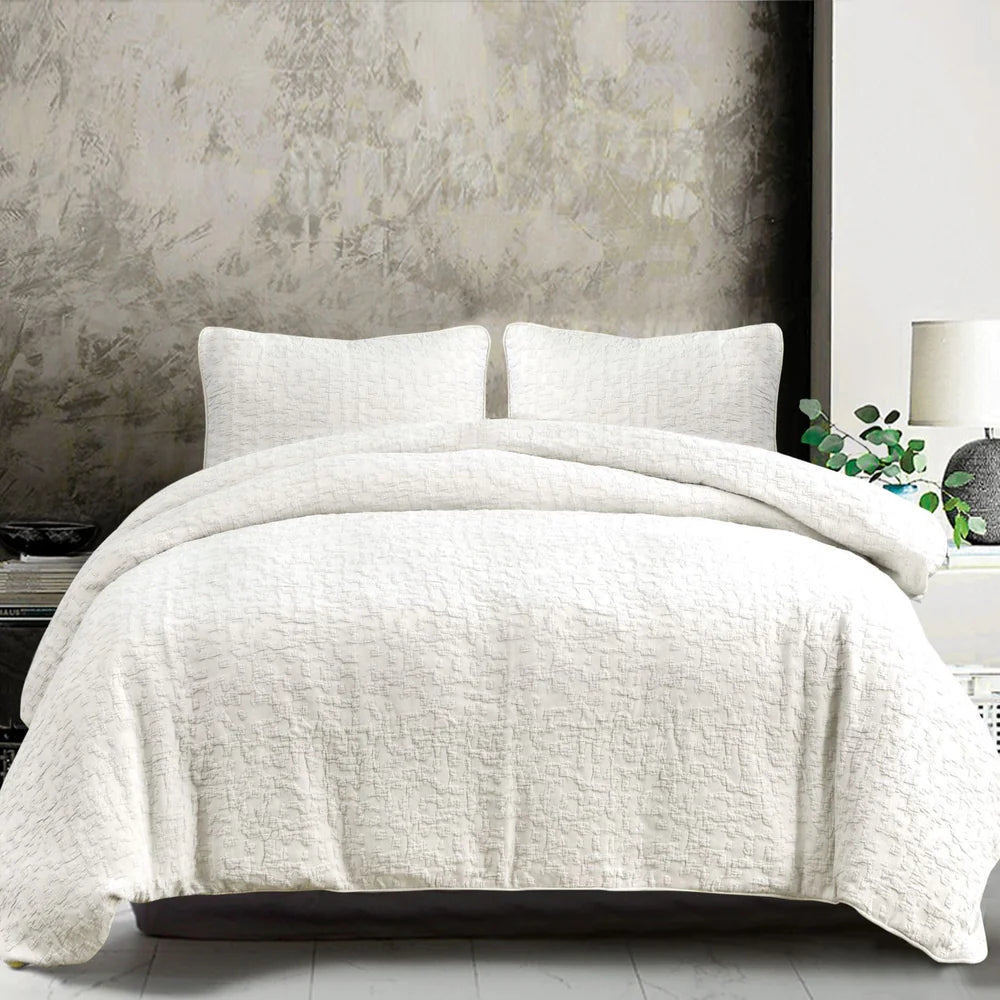 White quilted coverlet set on a bed with pillows against a textured wall.