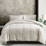 Textured beige quilt and pillow set on a bed with a neutral wall background