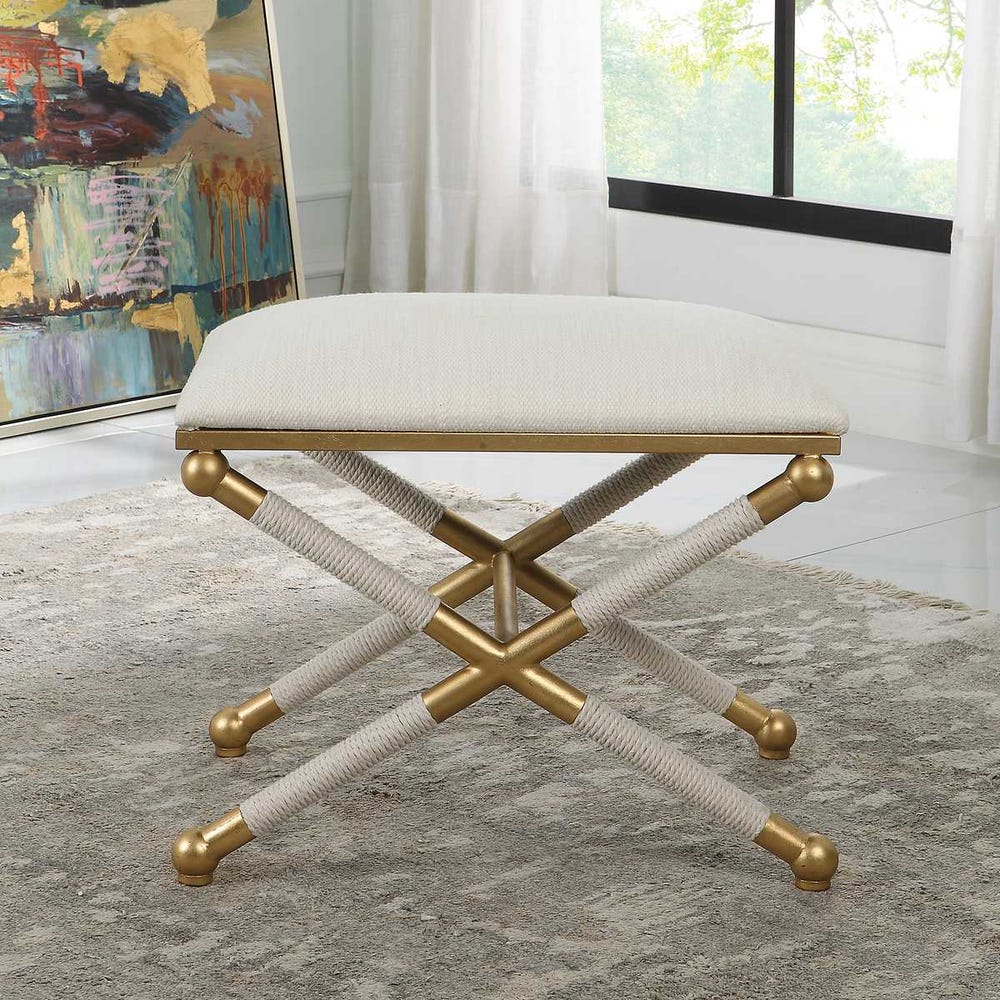 Decorative stool with gold legs and white cushion in a room with a painting and window.