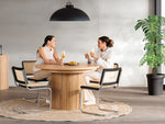 Two women sitting at a wooden dining table in a modern room with a plant and pendant light.