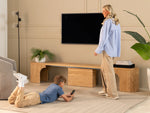 Woman standing and child lying on the floor in a living room with a TV and wooden furniture.