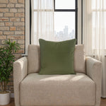 Beige armchair with a green pillow in a room with a stone wall and large windows.