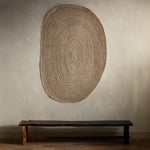 Woven wall art piece on a plain wall with a wooden bench below.