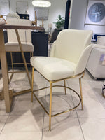 A white padded counter stool with gold-colored metal legs, displayed in a showroom setting.