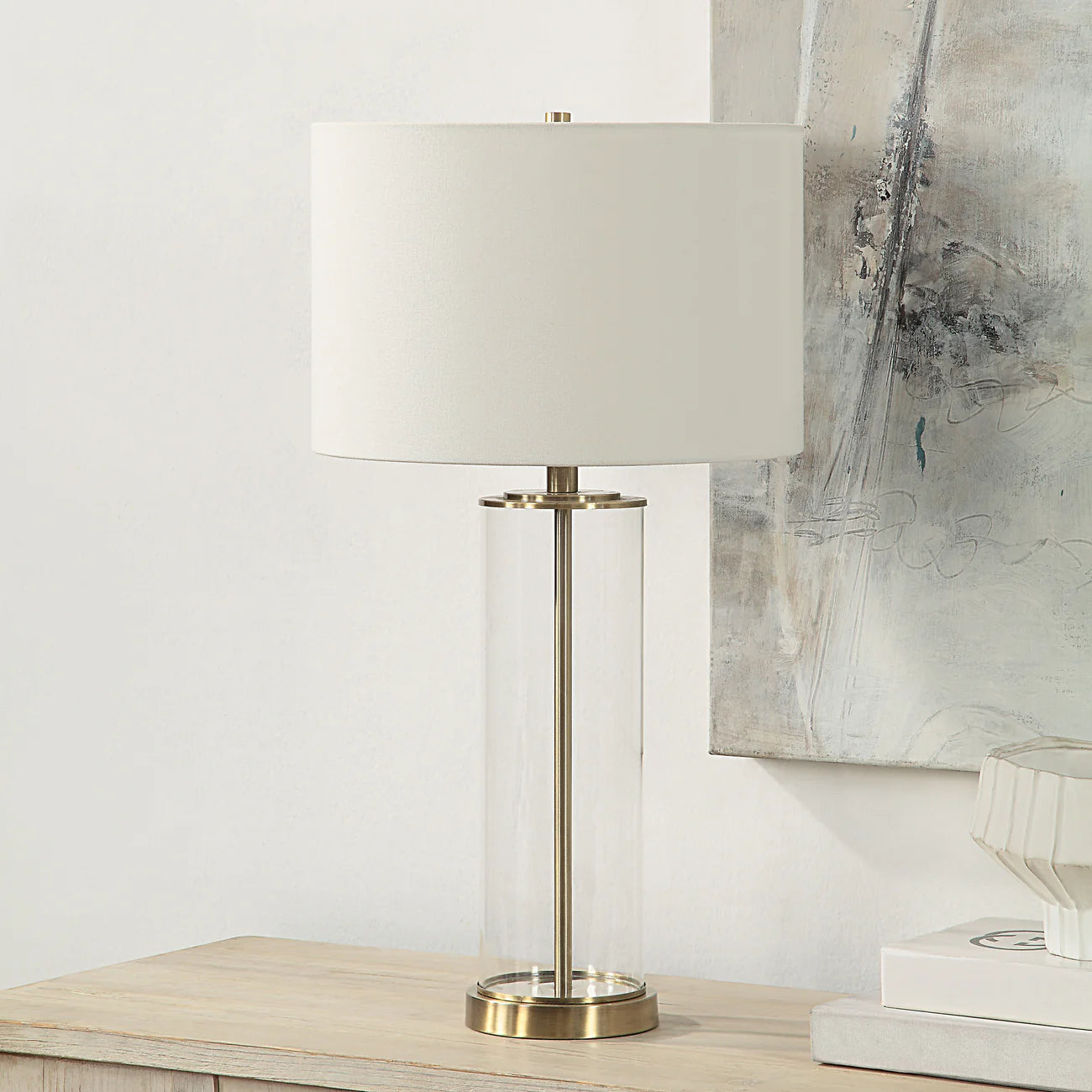 Table lamp with a white shade and brass base on a wooden surface, with abstract art in the background.