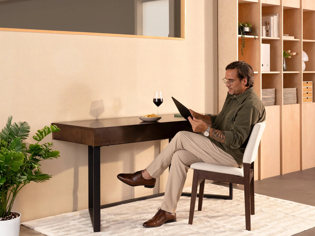 Man sitting at a desk reading a book with a glass of wine in a modern office setting.