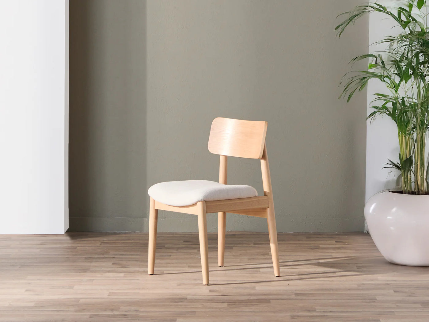 Wooden chair with a white cushion on a wooden floor against a gray wall with a plant in the corner.