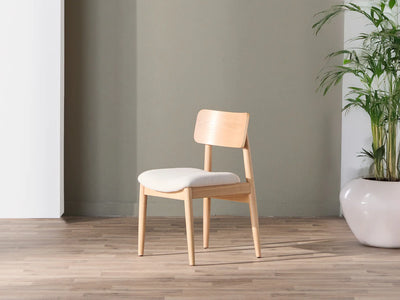 Wooden chair with a white cushion on a wooden floor against a gray wall with a plant in the corner.