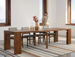 Dining room with a wooden table and chairs on a striped rug, person standing in the background.