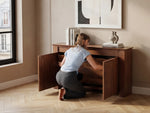Person opening a wooden cabinet in a room with a window and decorative items.