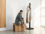 Woman sitting on a wooden bench next to a coat rack with a coat and handbag.