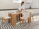 Woman setting a wooden dining table with white chairs in a modern kitchen.
