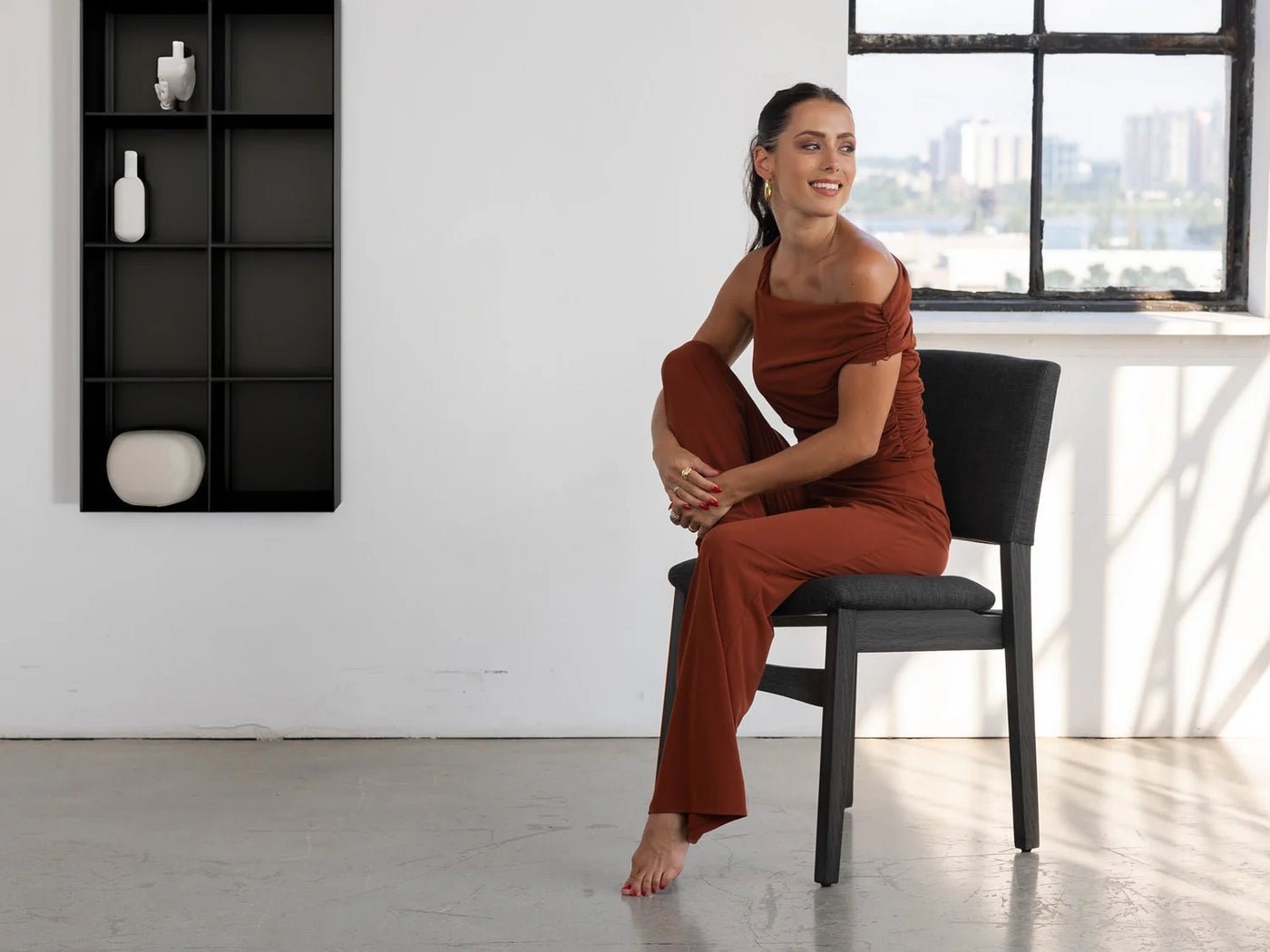 Woman in a rust-colored dress sitting on a black chair in a modern room with a cityscape view.