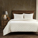 Bed with beige quilt and pillows in a bedroom setting with wooden nightstand and lamp.