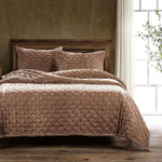 Quilted brown bedspread on a wooden bed with pillows in a room with a window and plant.