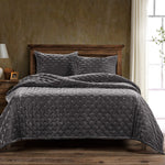 Gray quilted coverlet on a bed with wooden headboard and side tables in a room with a textured wall and window.