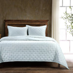 Light blue quilted coverlet on a bed with wooden headboard and pillows, in a room with a window and plant.