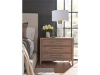 Wooden nightstand with lamp and flowers next to a bed in a bedroom.
