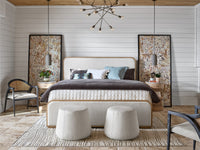 A styled bedroom interior featuring a bed with a cream upholstered headboard and footboard, layered bedding in dark brown and light blue, two matching cylindrical ottomans at the foot of the bed, and two tall framed botanical wall art pieces against white shiplap walls.