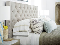 A styled bedroom scene featuring a large tufted upholstered headboard in a light beige fabric, dressed with white bedding, patterned accent pillows, and a textured quilt.
