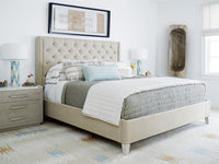 A beige upholstered bed with a tufted headboard, styled with gray and white bedding, a light blue accent pillow, a gray nightstand, and a patterned area rug in a bright bedroom.