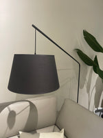 A black floor lamp with a modern design, hung in a room with a white sofa and a green potted plant in the background.