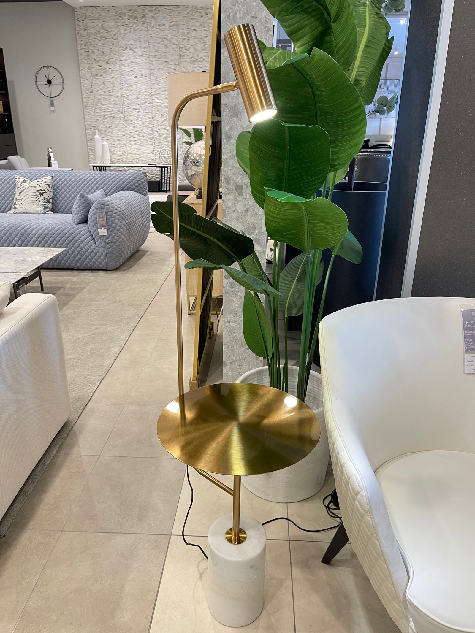 A gold-colored accent table with a marble base and a cylindrical shape, standing in a room with a large green plant and a cozy seating setup.